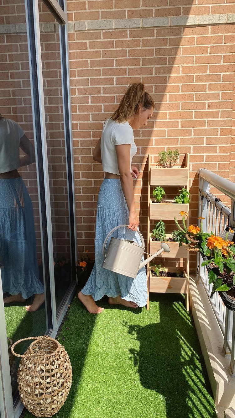 A woman watering small plants in a wooden vertical planter on a sunny apartment balcony.