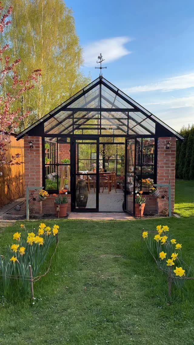 21 Backyard Landscape Design Ideas for Outdoor Living 18 A glass and brick greenhouse with a black frame stands on a green lawn with yellow flowers.