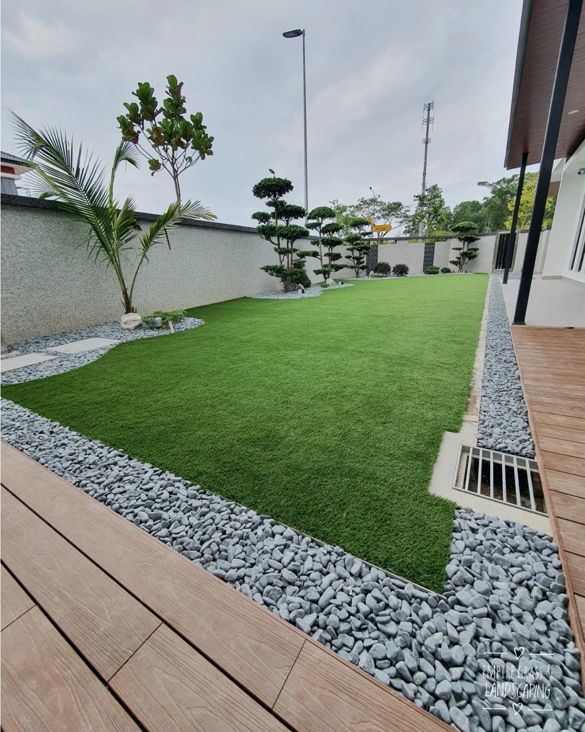 21 Backyard Landscape Design Ideas for Outdoor Living 16 Artificial turf lawn bordered by grey river stones next to a wooden deck and white wall.