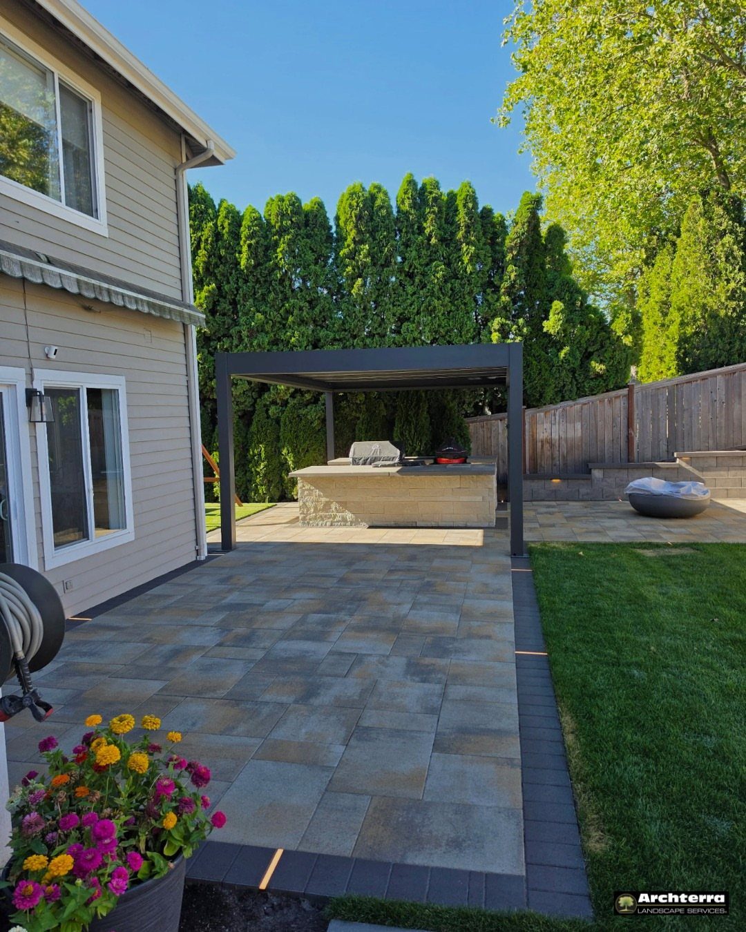 21 Backyard Landscape Design Ideas for Outdoor Living 2 Stone patio pavers leading toward a dark pergola and an outdoor kitchen area.