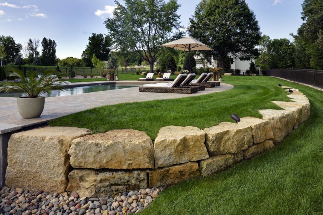 21 Backyard Landscape Design Ideas for Outdoor Living 12 A curved natural stone retaining wall bordering a lush green lawn and a swimming pool area.