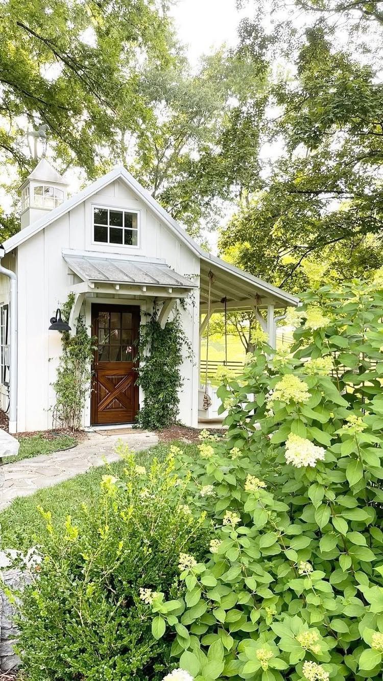 21 Backyard Landscape Design Ideas for Outdoor Living 17 A small white wooden garden shed with a dark wood door surrounded by green plants.