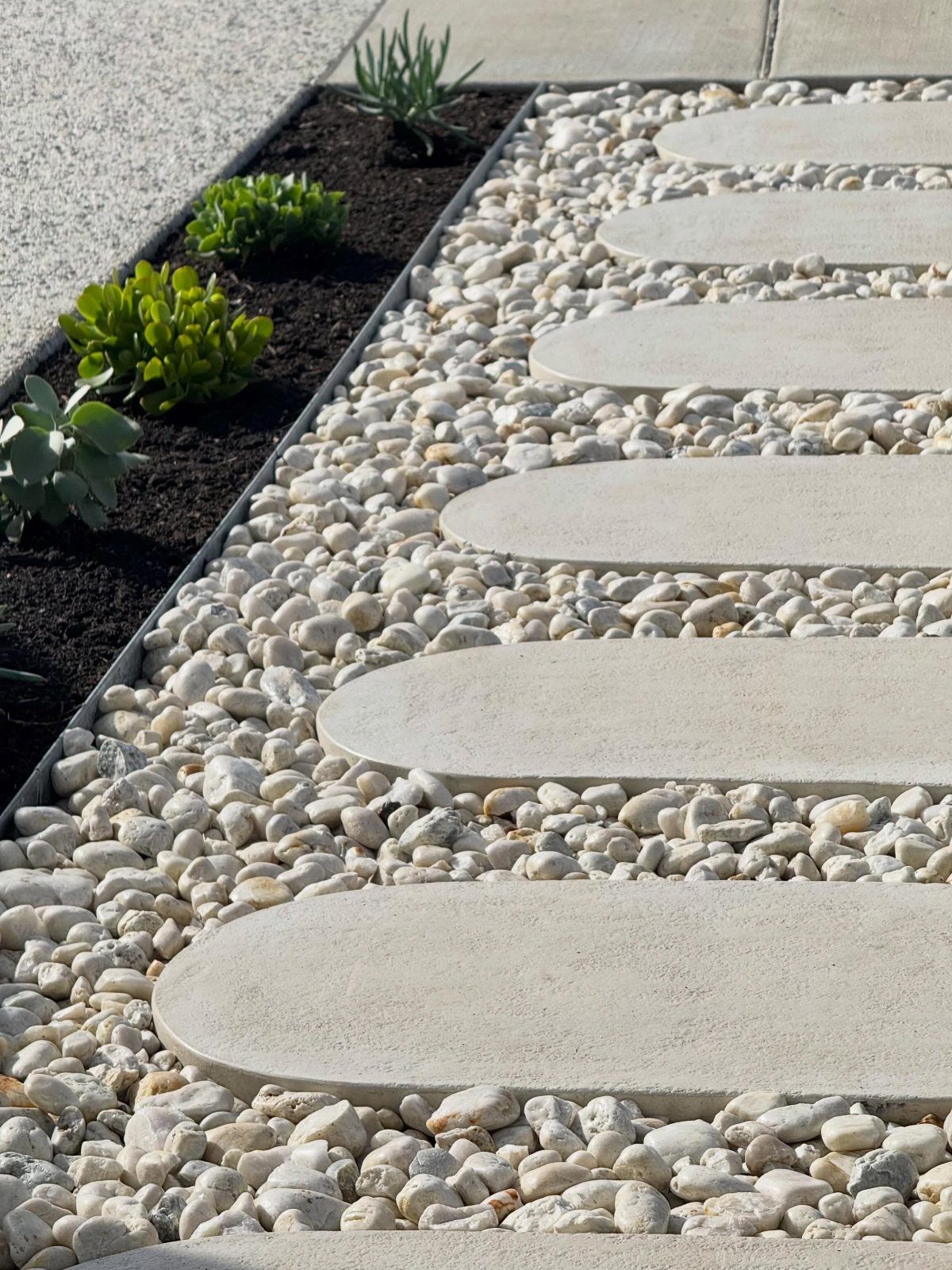 21 Backyard Landscape Design Ideas for Outdoor Living 21 Oval light grey stepping stones embedded in white pebbles next to a small succulent garden bed.
