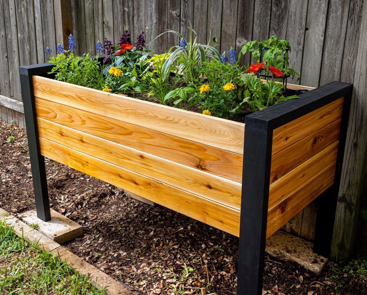 An elevated wooden planter box with black trim filled with colorful flowers against a fence.
