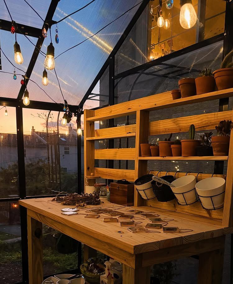 String lights glow inside a greenhouse featuring a wooden workbench and potted plants at sunset.