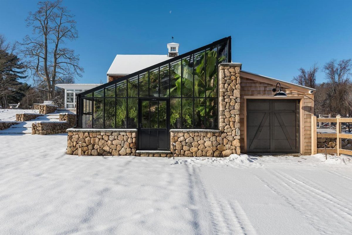 A modern glass lean-to greenhouse attached to a stone and wood building in a snowy landscape.