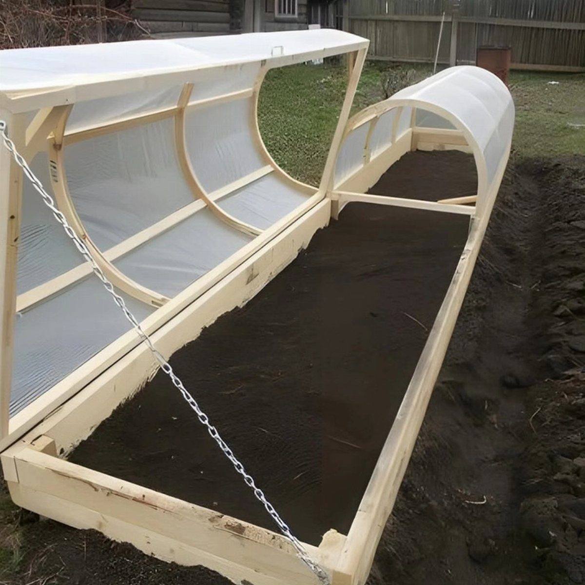 A wooden raised garden bed with a white plastic hoop house structure over dark soil.