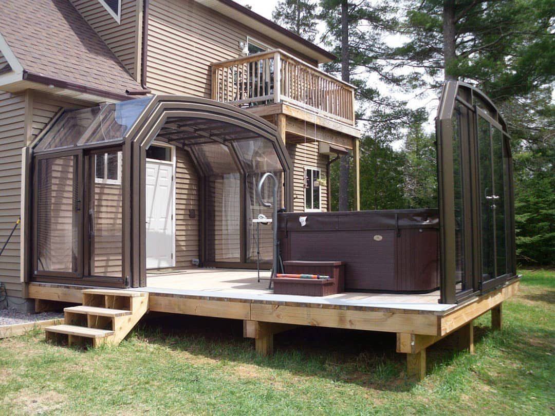 7 Covered Hot Tub Ideas for Backyard Privacy and Style (2026) 3 A brown hot tub on a wooden deck under a curved glass enclosure attached to a house.