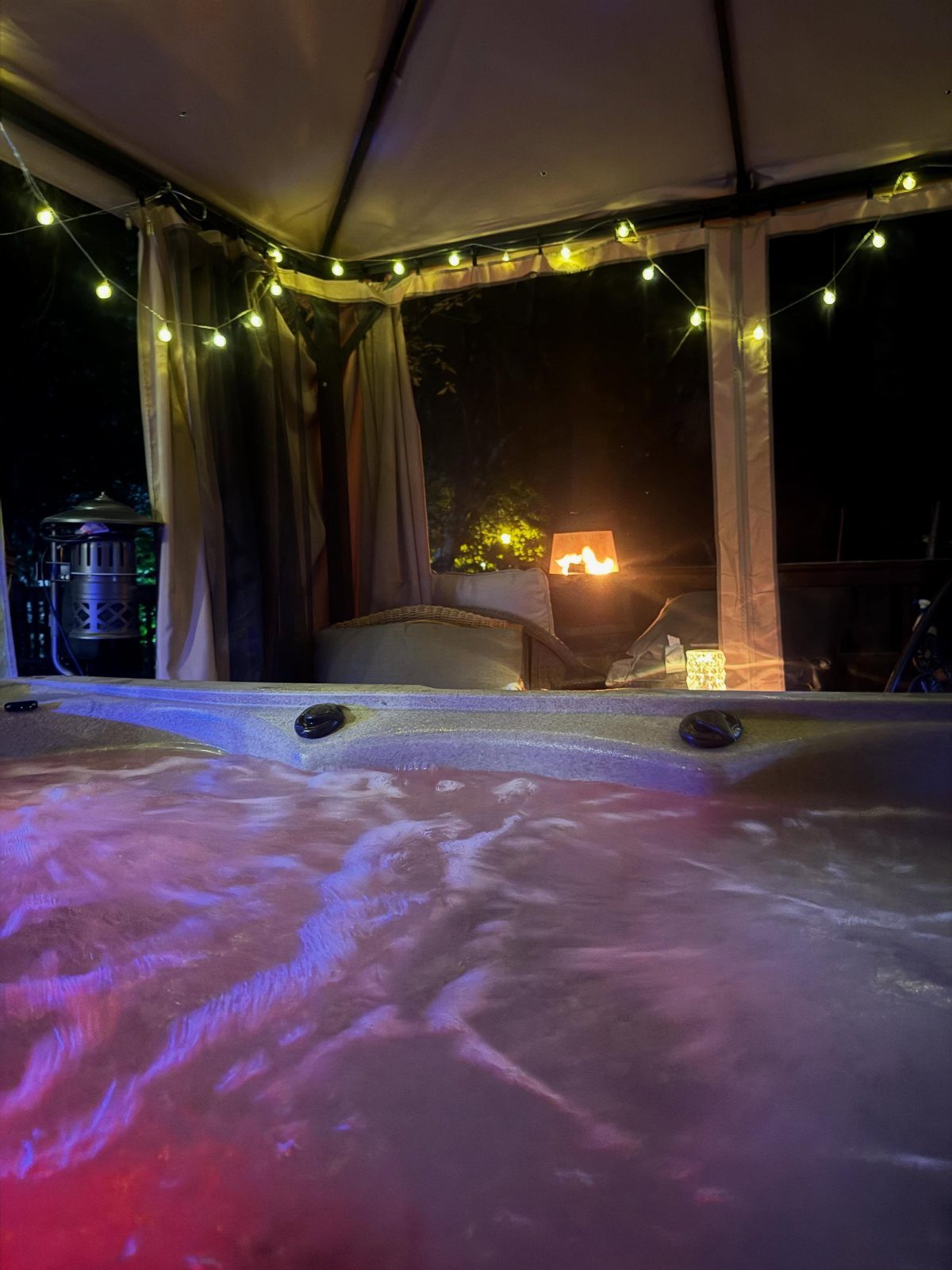 7 Covered Hot Tub Ideas for Backyard Privacy and Style (2026) 1 Bubbling hot tub water with colorful lights under a canopy decorated with glowing string lights.