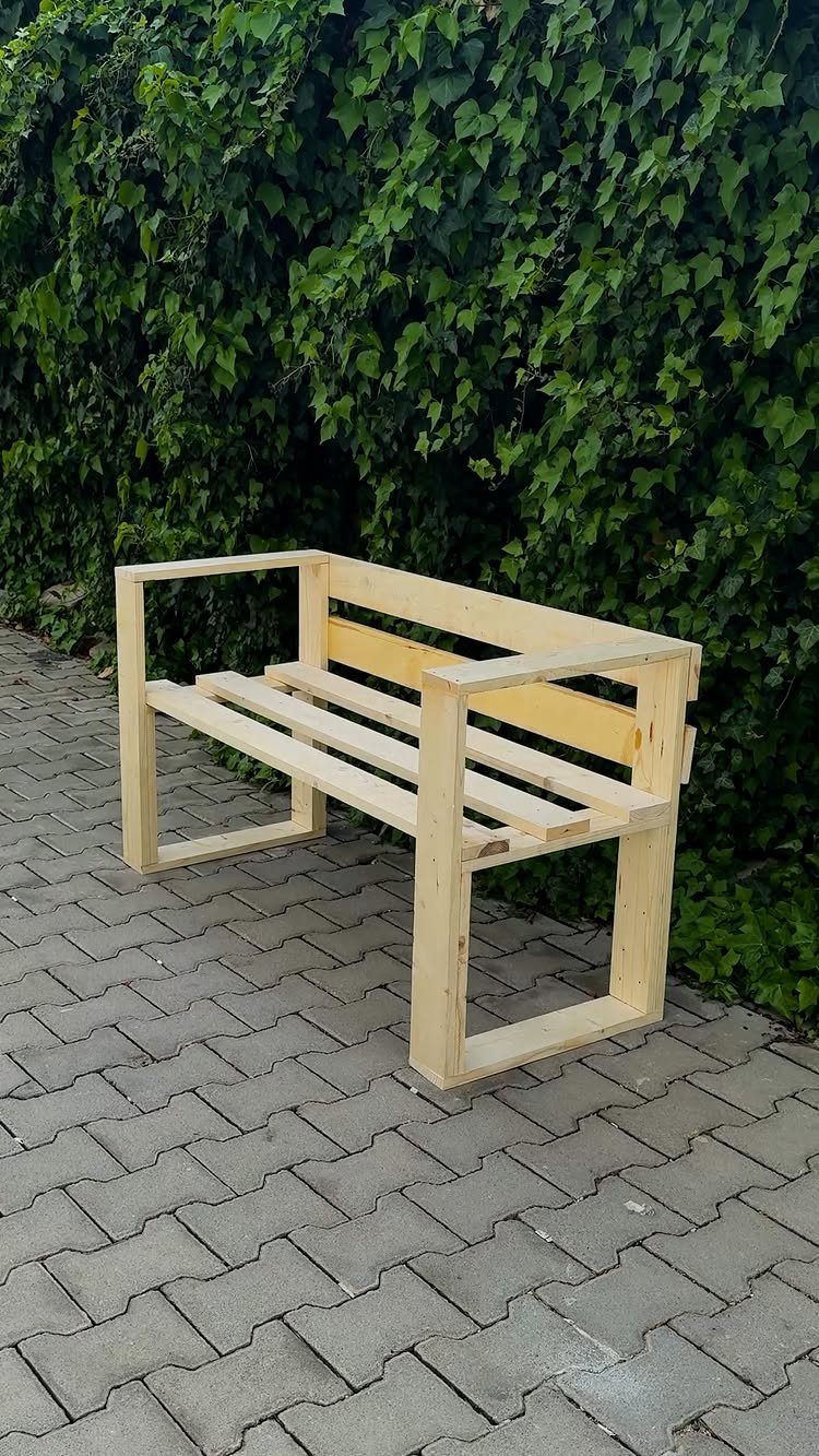 DIY Garden Bench: 8 Plans from Classic Backrests to Cinder Block Builds 3 A light wooden slatted garden bench sits on a stone patio against an ivy wall.