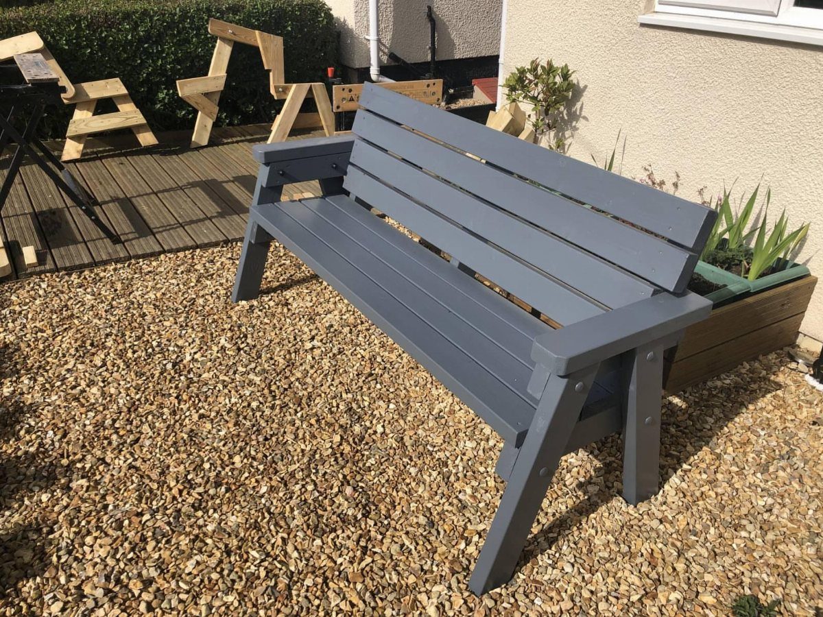 DIY Garden Bench: 8 Plans from Classic Backrests to Cinder Block Builds 1 A grey wooden garden bench with a slatted backrest resting on a gravel patio.