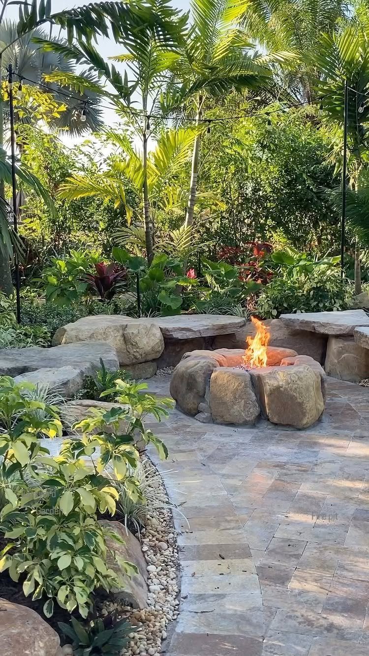 12 Dream Backyard Garden Ideas with Outdoor Kitchens to Natural Pools 2 A stone fire pit with a burning flame on a paved patio surrounded by tropical plants.