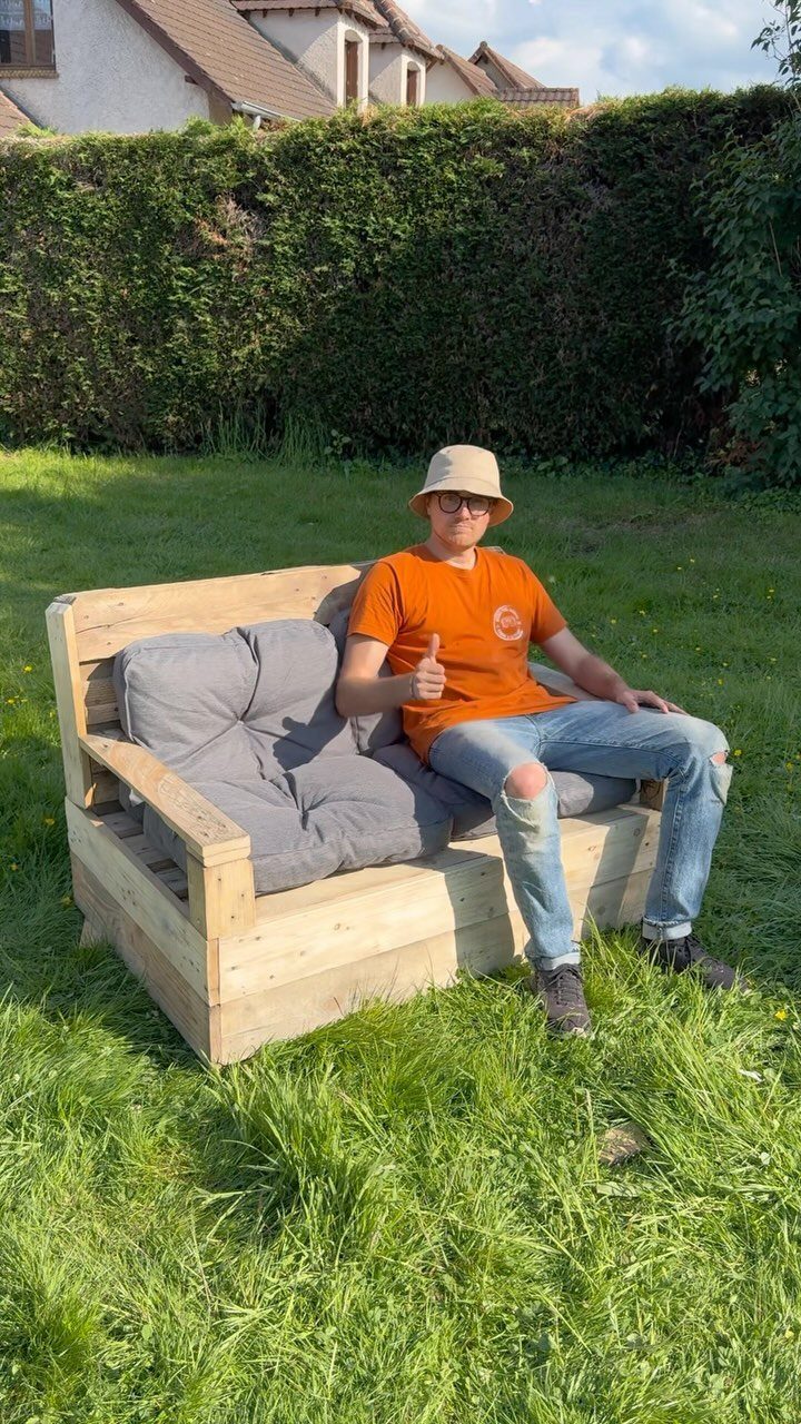 7 Easy DIY Patio Ideas With Paver Floors, Pallets and Gravel 3 A person sitting on a DIY pallet sofa with grey cushions on a green lawn.