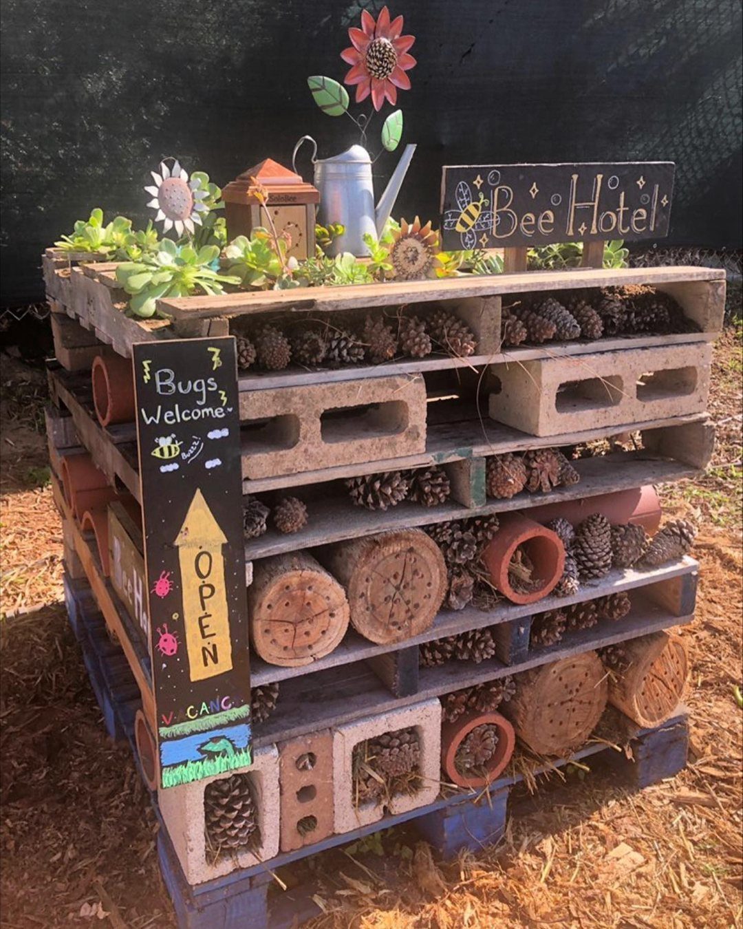 A multi-layered wooden pallet bug hotel filled with pinecones, logs, and terracotta pots.