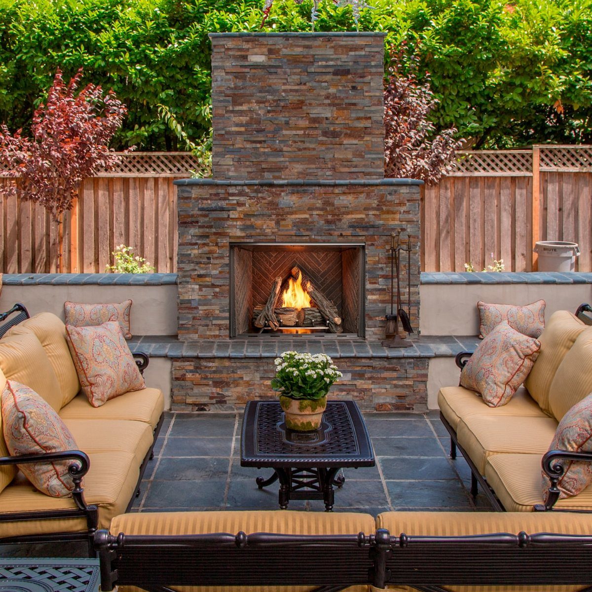 Stone outdoor fireplace with a burning fire surrounded by comfortable patio sofas and cushions.