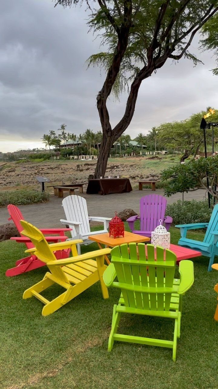 Colorful Adirondack chairs in various bright hues arranged on a green lawn under a large tree.