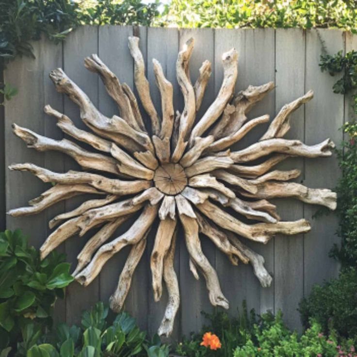 Driftwood sunburst wall art mounted on a grey wooden fence surrounded by green garden plants.