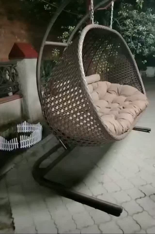 A woven wicker hanging egg chair with beige cushions on a metal stand outdoors.