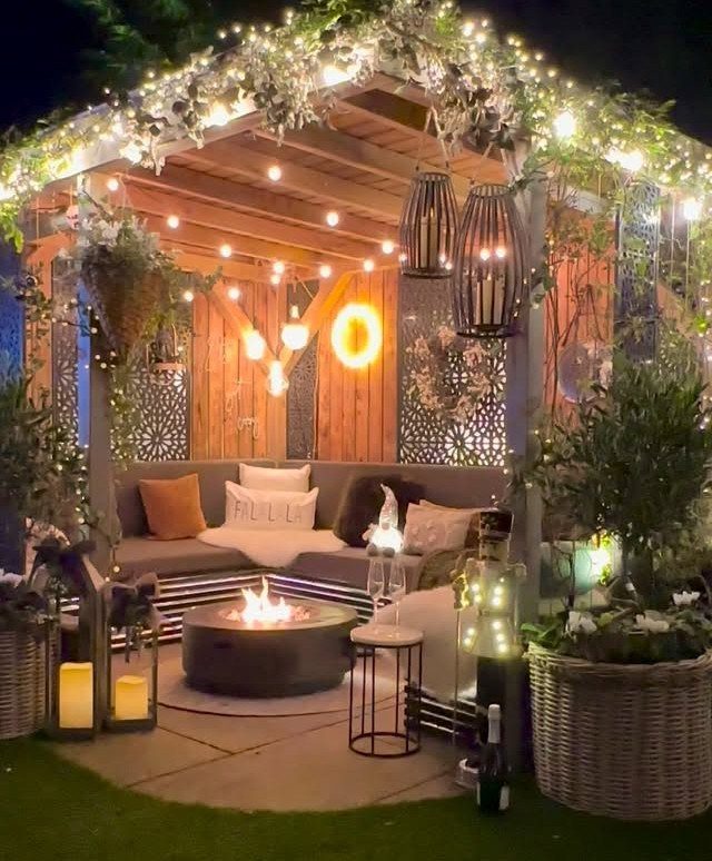 A wooden pergola decorated with fairy lights, greenery, and a cozy seating area with a fire pit.