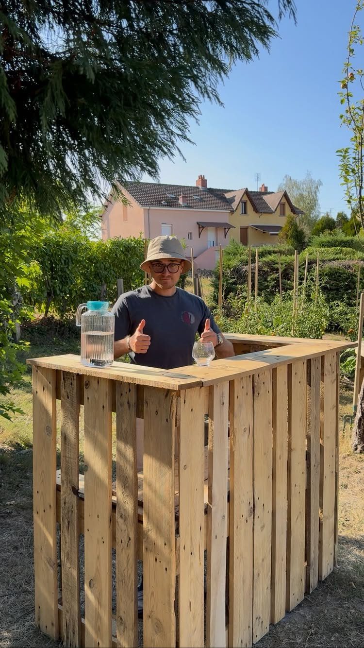 A man standing behind a tall outdoor bar constructed from light wooden pallets in a garden.