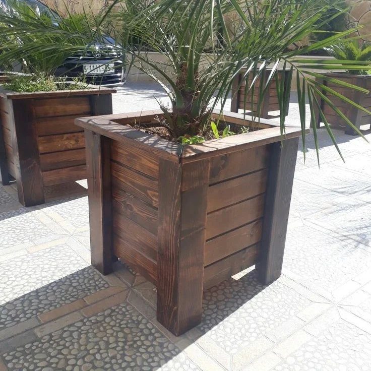 Dark stained wooden square planters with green palm plants on a stone patio.
