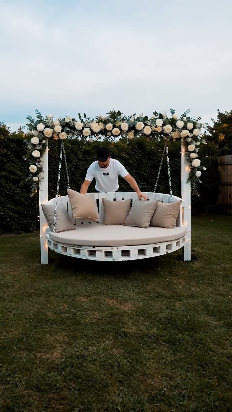 A man leaning over a large, circular white pallet swing decorated with flowers on a lawn.