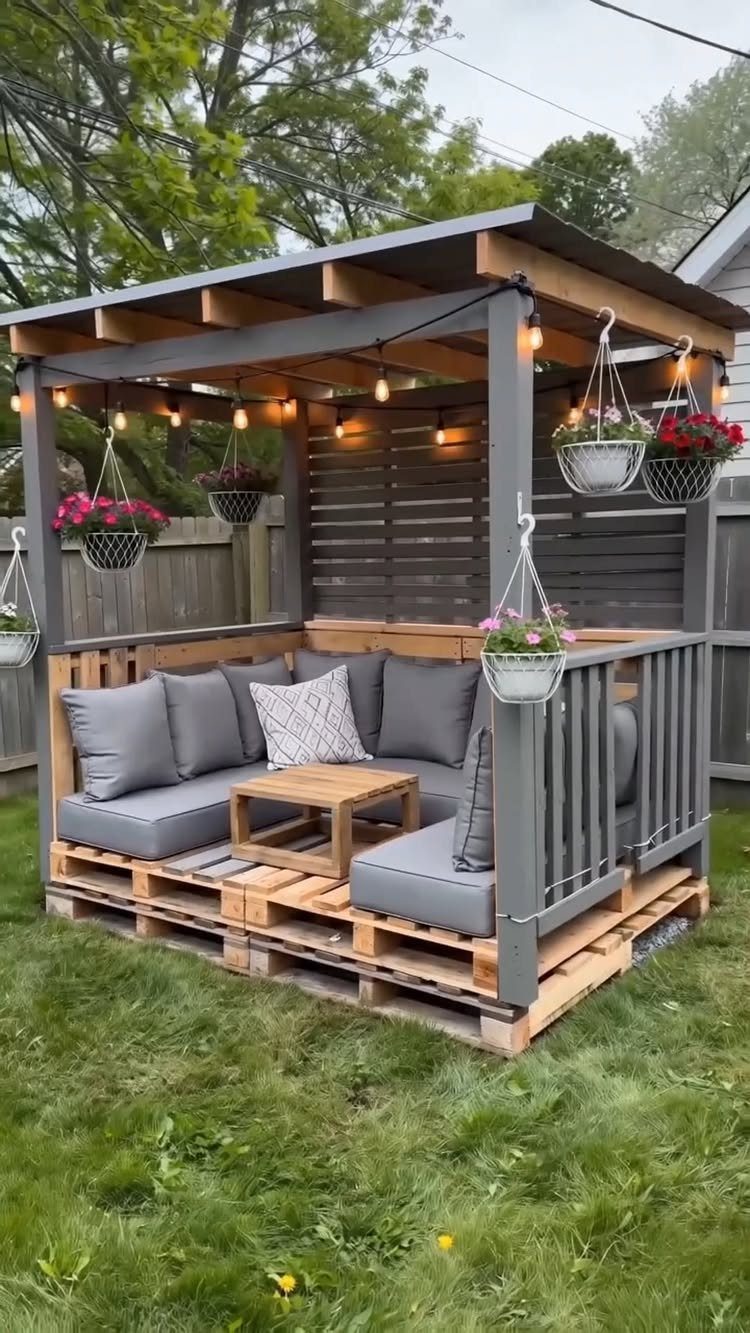A DIY pallet lounge with grey cushions and string lights under a wooden pergola structure.