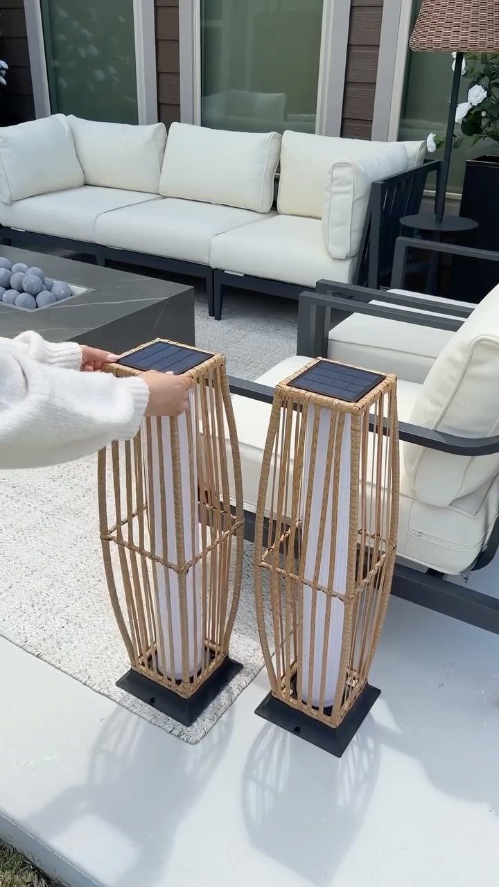 Two tall rattan-style solar lanterns sitting on a light patio near white outdoor lounge furniture.