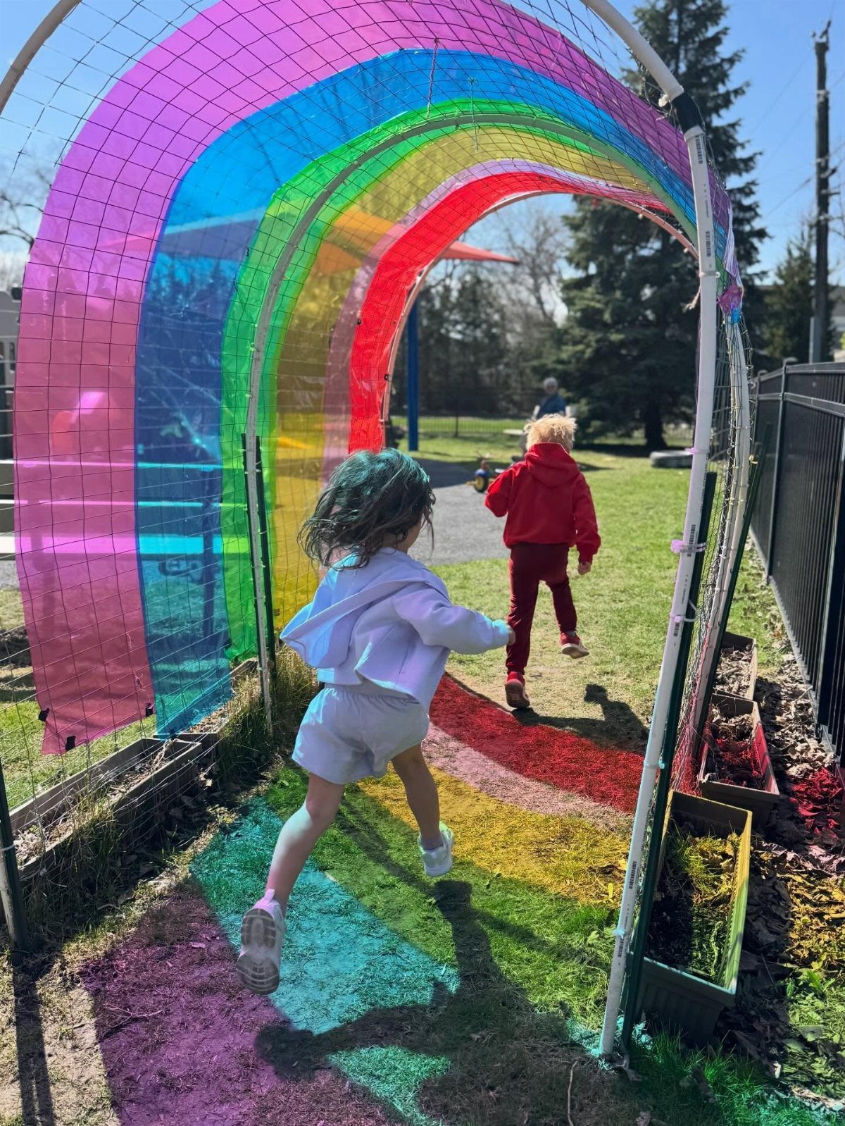 Two children running through a bright rainbow-colored mesh tunnel in an outdoor play area.