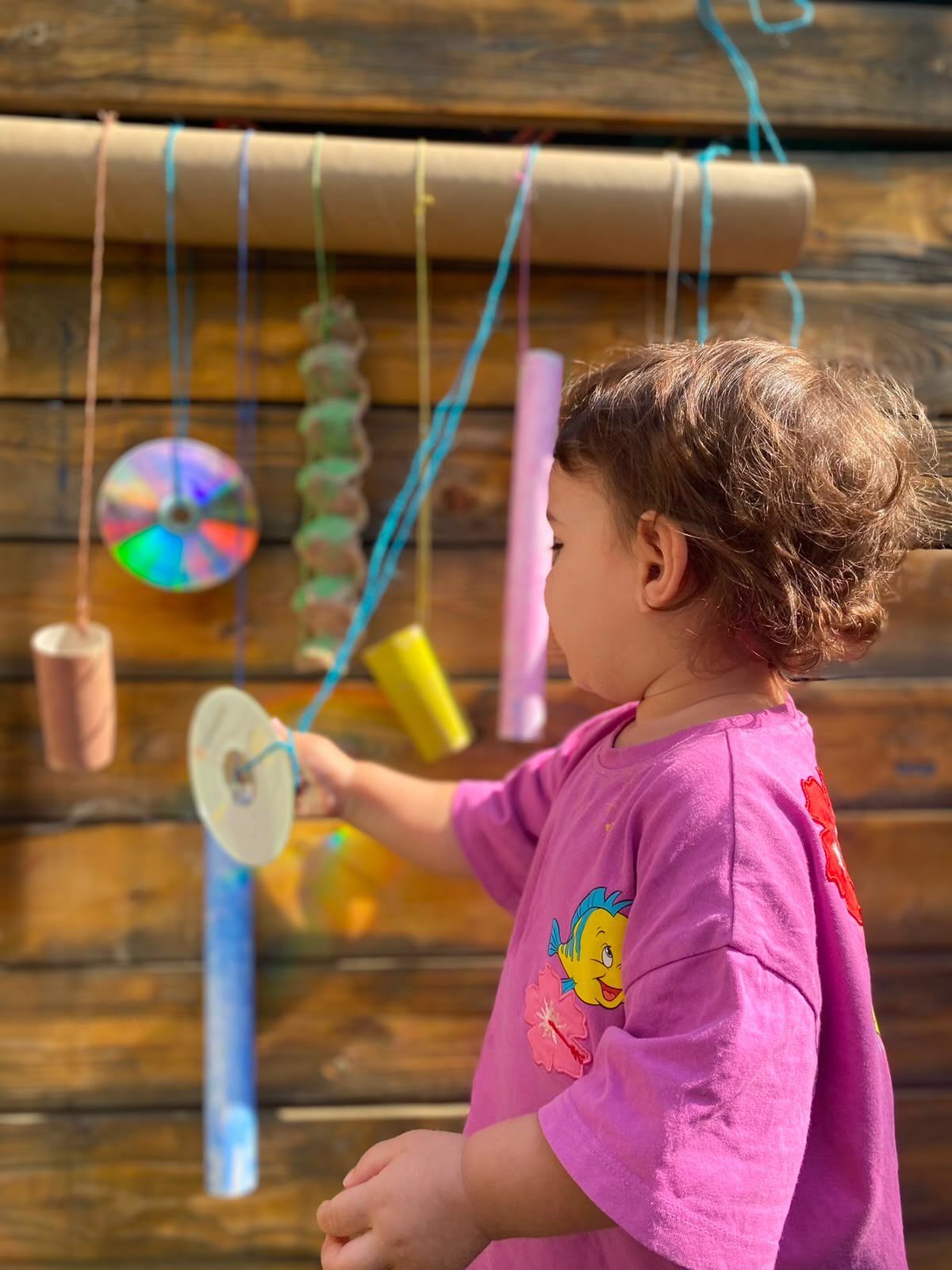 A young child reaches for colorful hanging objects suspended from a wooden beam outdoors.