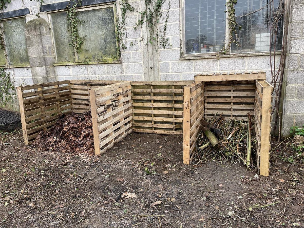 Wooden pallet compost bins filled with leaves and plant debris against a cinder block wall.