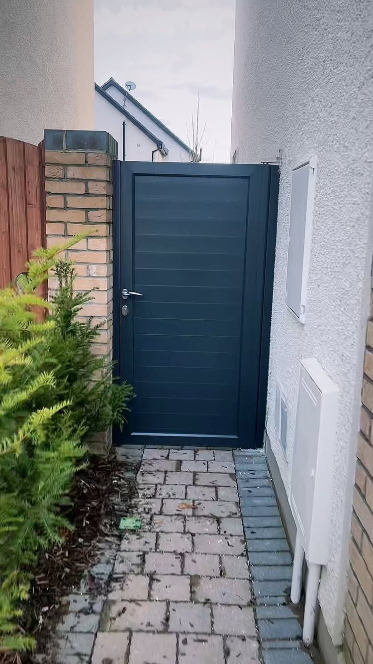 A dark grey modern garden gate set between a brick pillar and a white wall.