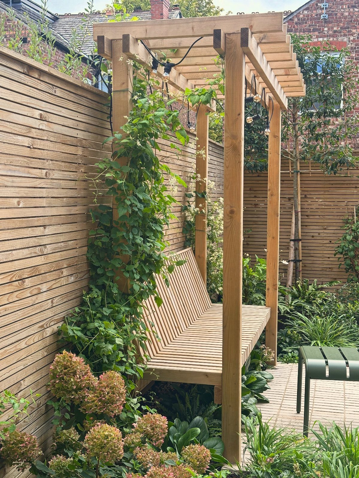 Green climbing plants growing up the wooden posts of a garden pergola with a built-in bench.