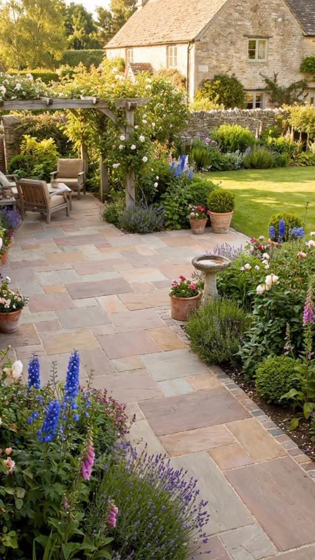 Stone patio surrounded by lush, colorful flowering plants and greenery near a stone house.
