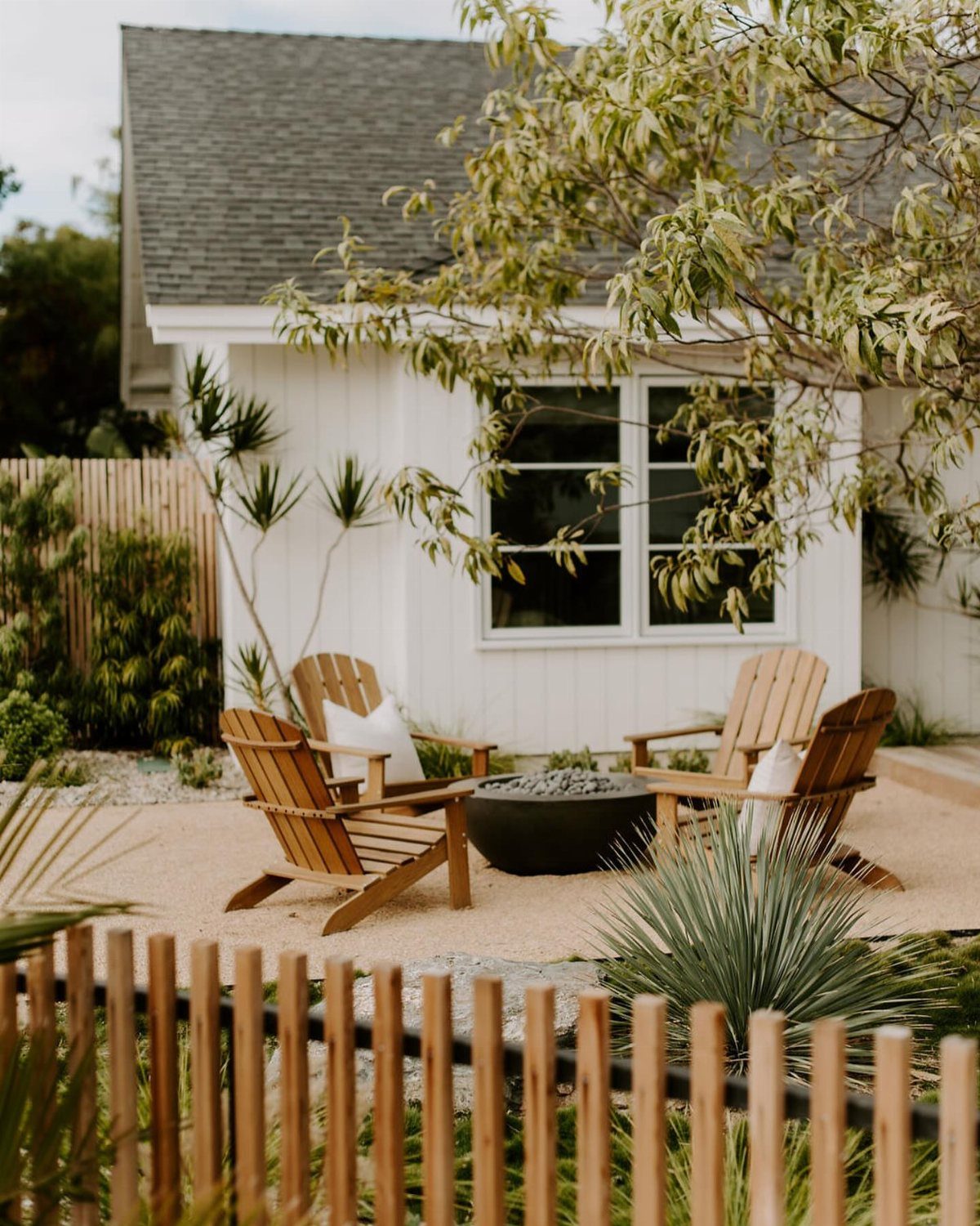 Wooden Adirondack chairs arranged around a black fire pit on a gravel patio near a white house.