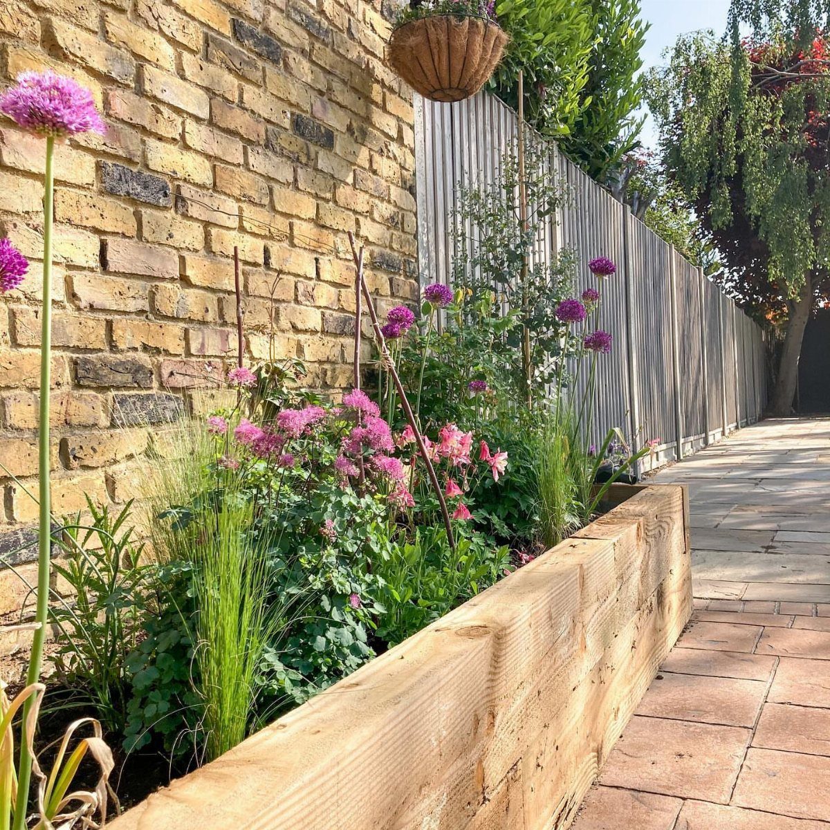 A wooden raised garden border filled with pink flowers and green plants next to a brick wall.