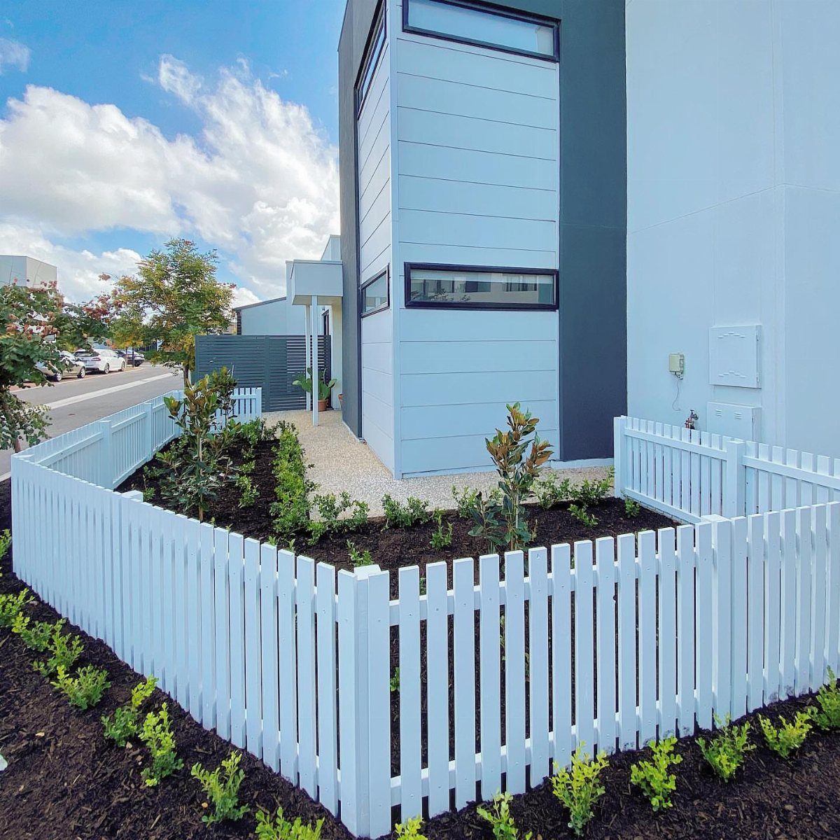 17 Small Front Yard Landscape Ideas with Photo Inspiration 10 A white picket fence surrounds a small landscaped garden bed in front of a modern house.