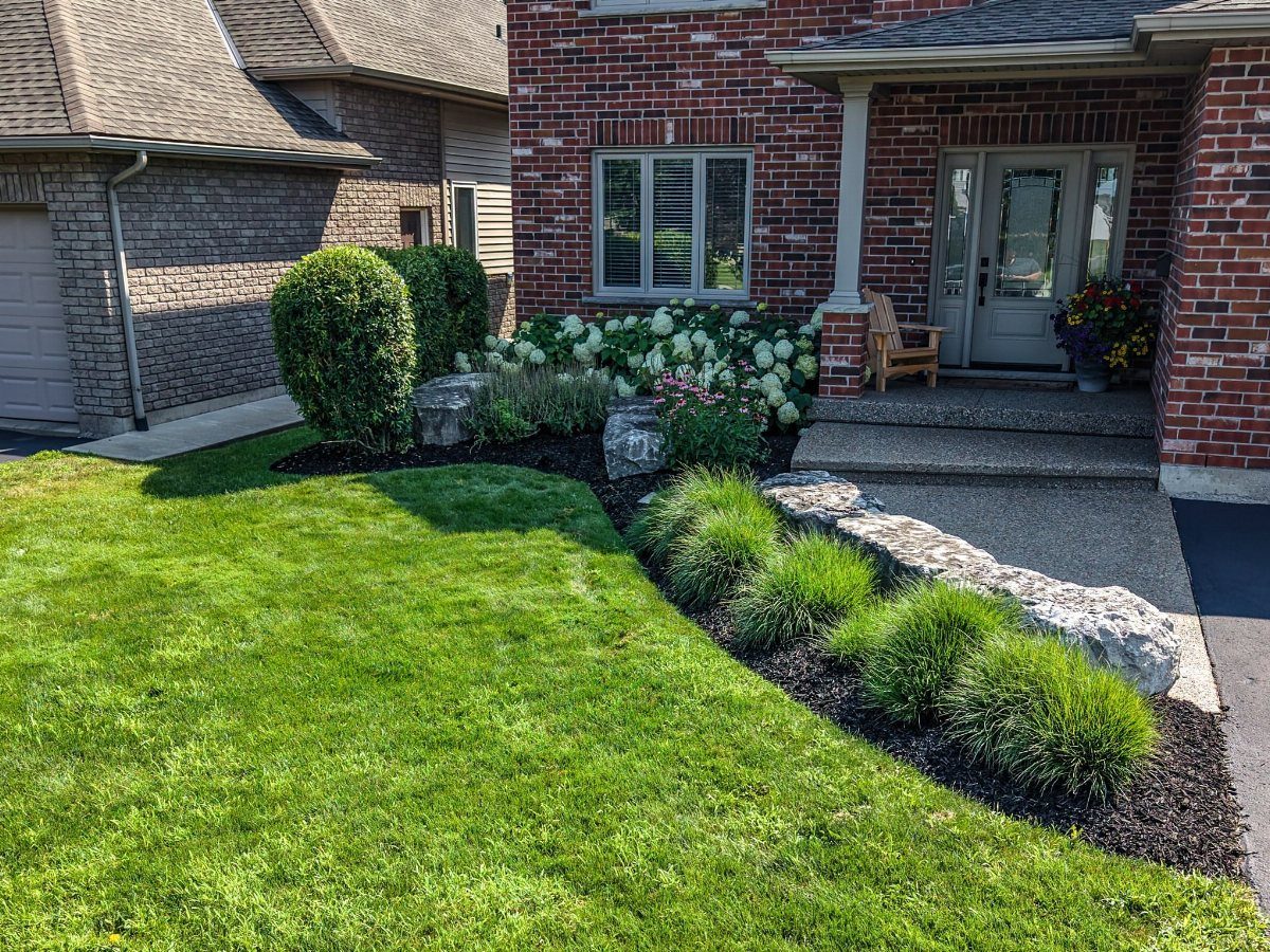 17 Small Front Yard Landscape Ideas with Photo Inspiration 2 A landscaped front yard with dark mulch, green shrubs, and a stone path near a brick house.