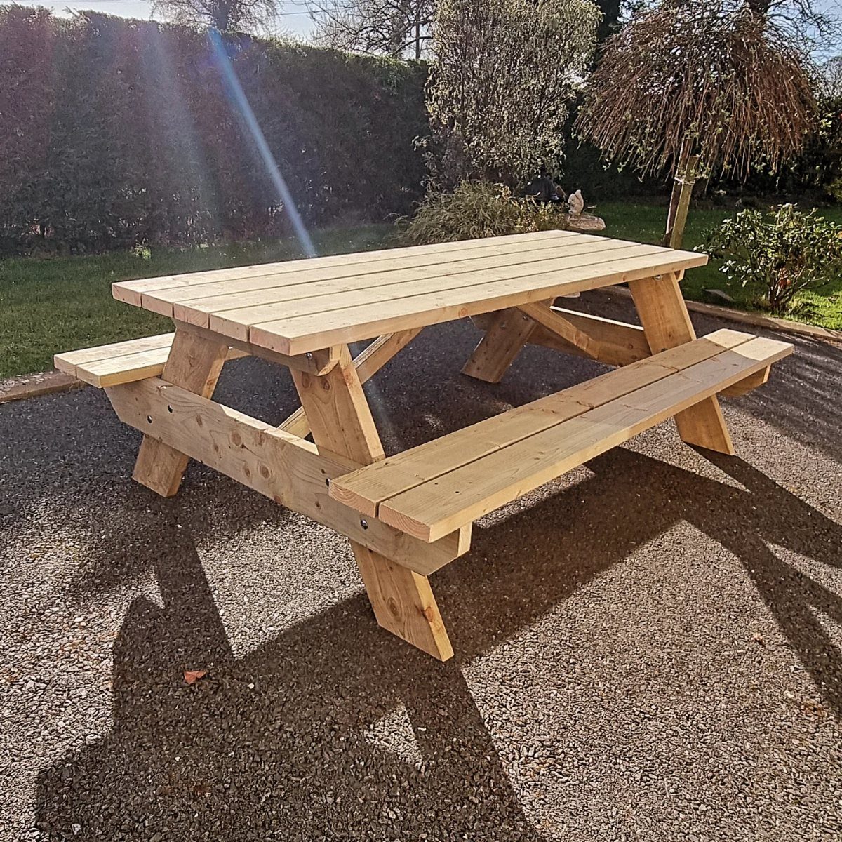 A wooden picnic table with attached benches on a gravel patio in a garden.