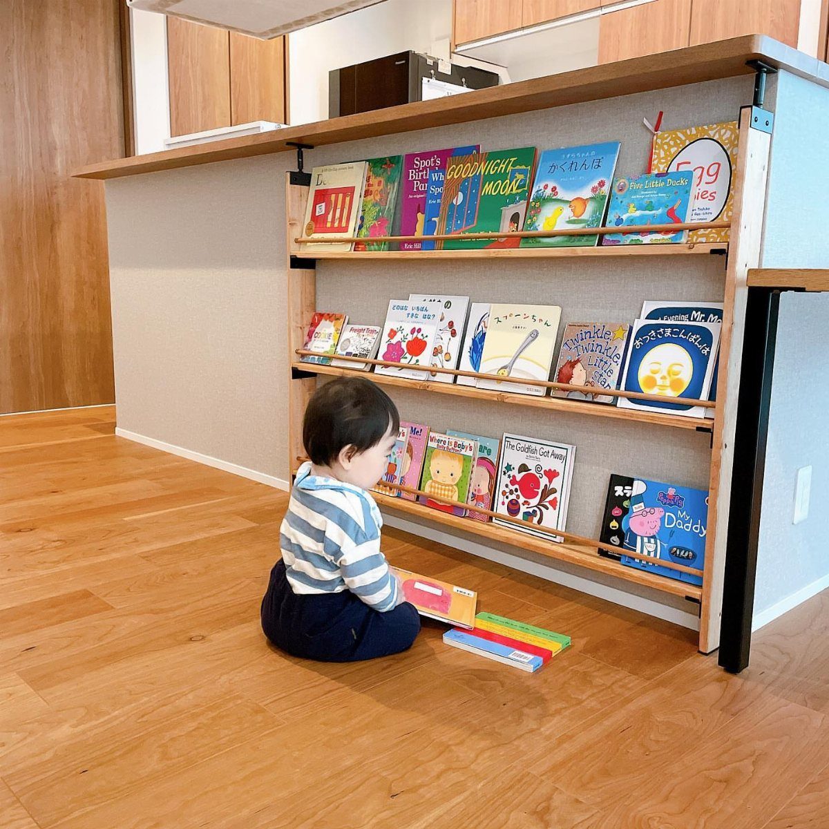 A toddler sits on a wooden floor playing with books in front of a low wooden bookshelf.