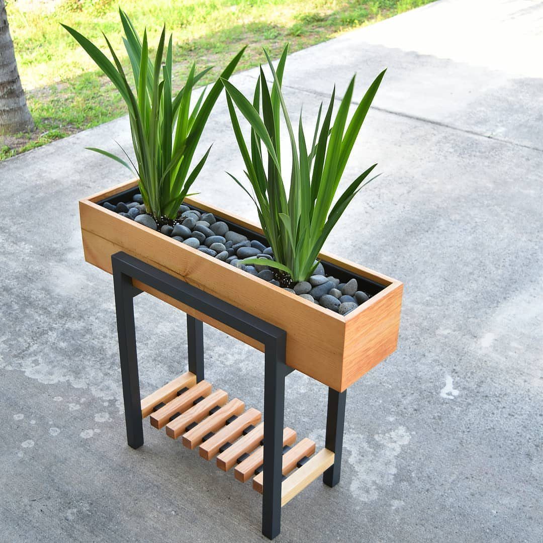 A rectangular wooden planter with green plants and black stones on a metal stand.