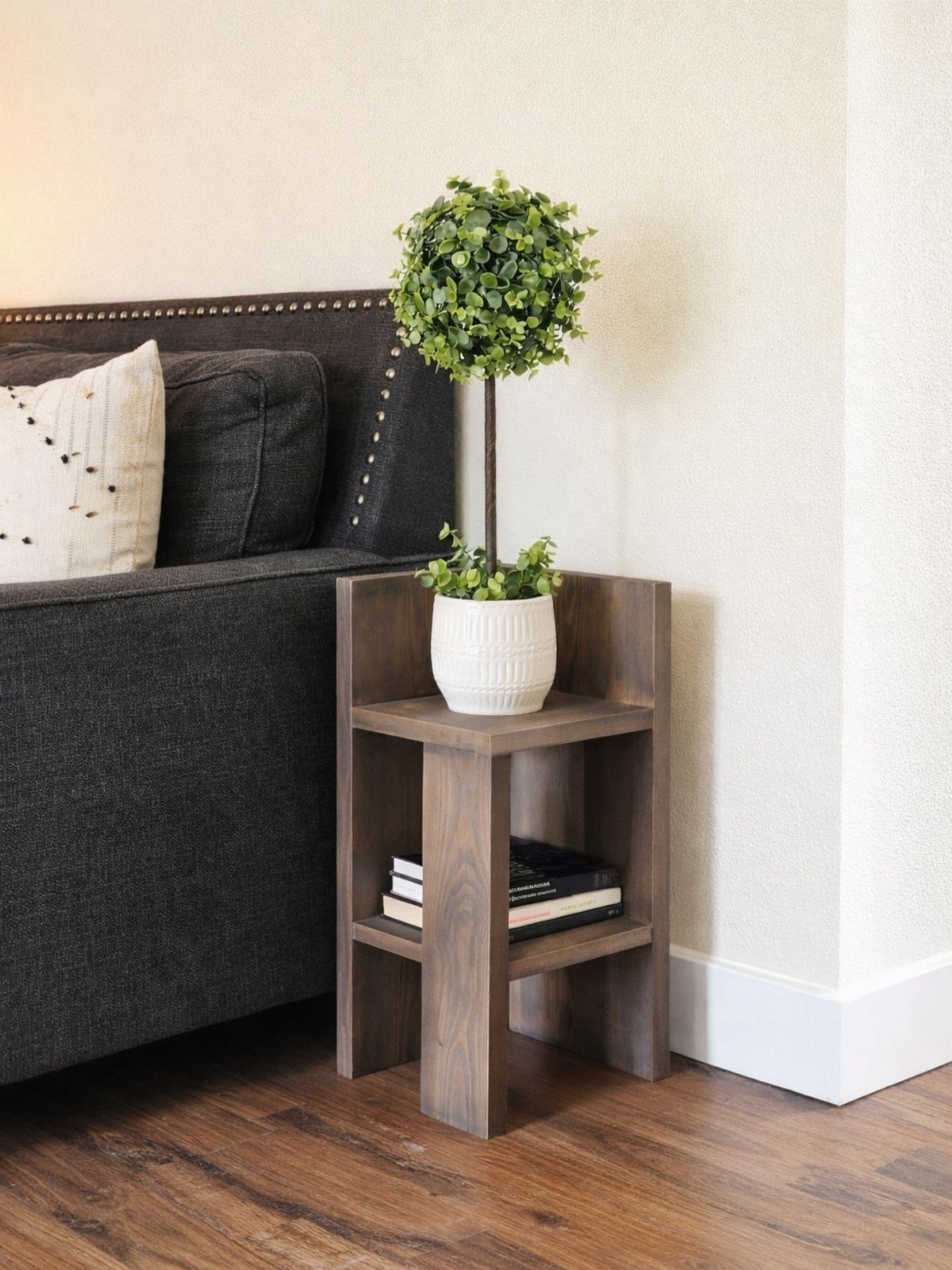A dark wood corner side table with two shelves holding a plant and books.