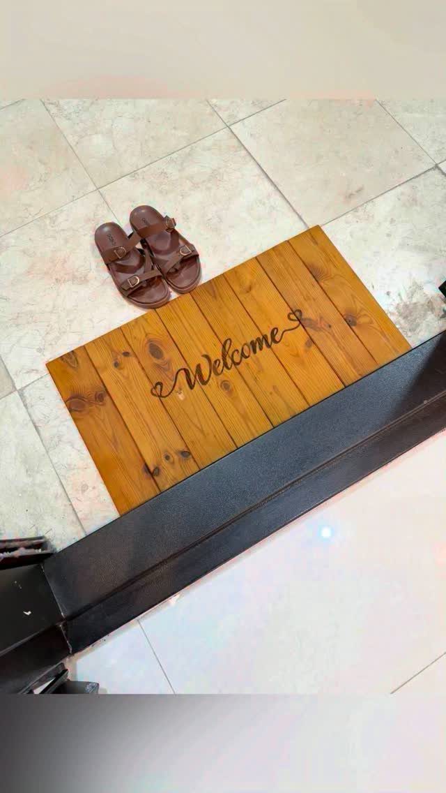 A wooden welcome doormat on a tiled floor with a pair of brown sandals.