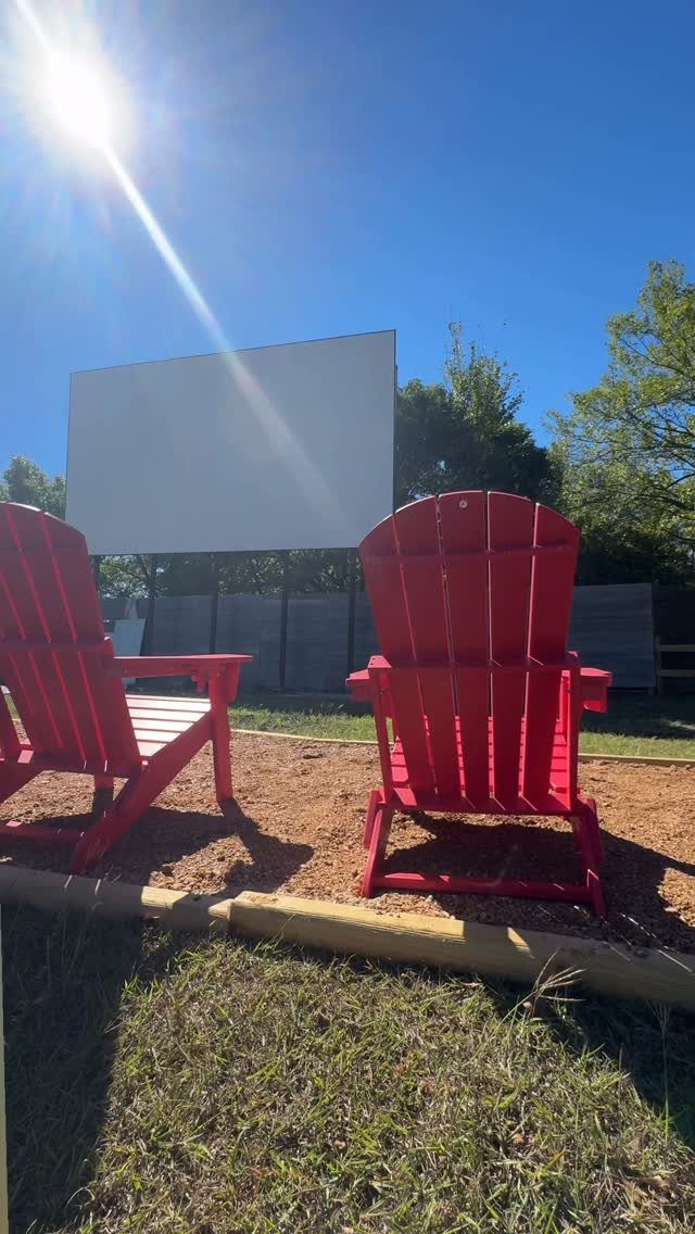 Two red Adirondack chairs facing a large white screen outdoors under bright sunlight.