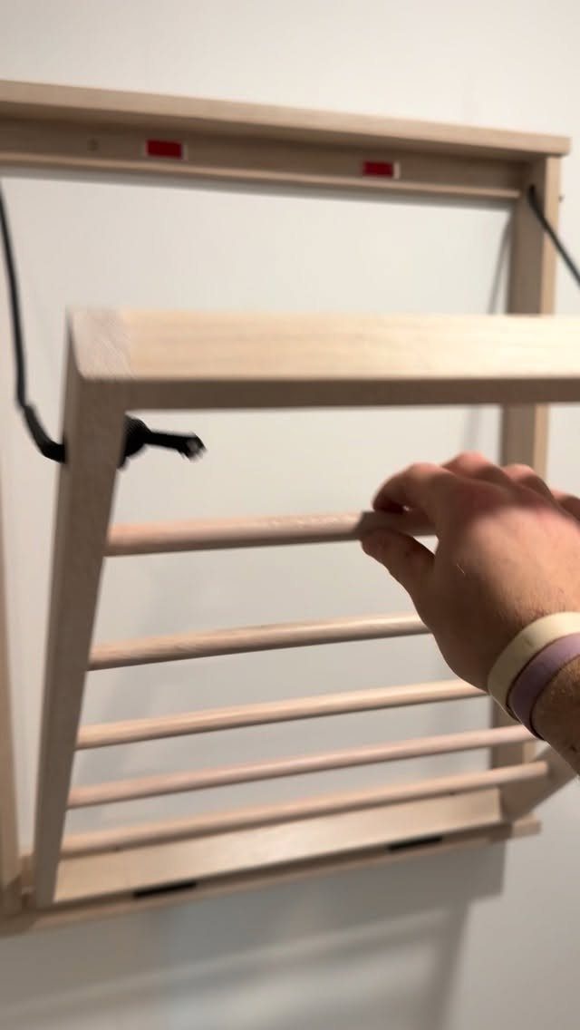 A wooden folding clothes drying rack mounted on a white wall being touched by a hand.