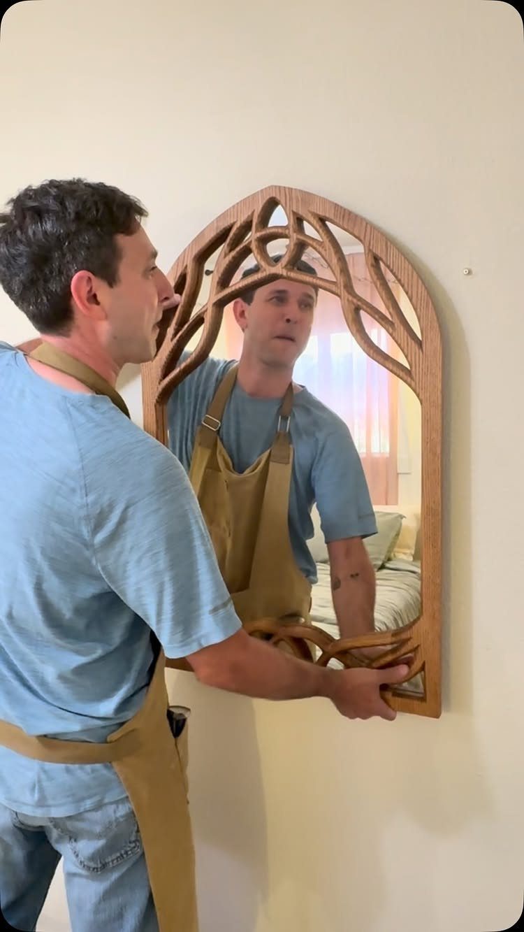 A man holding an intricately carved arched wooden mirror frame against a light-colored wall.