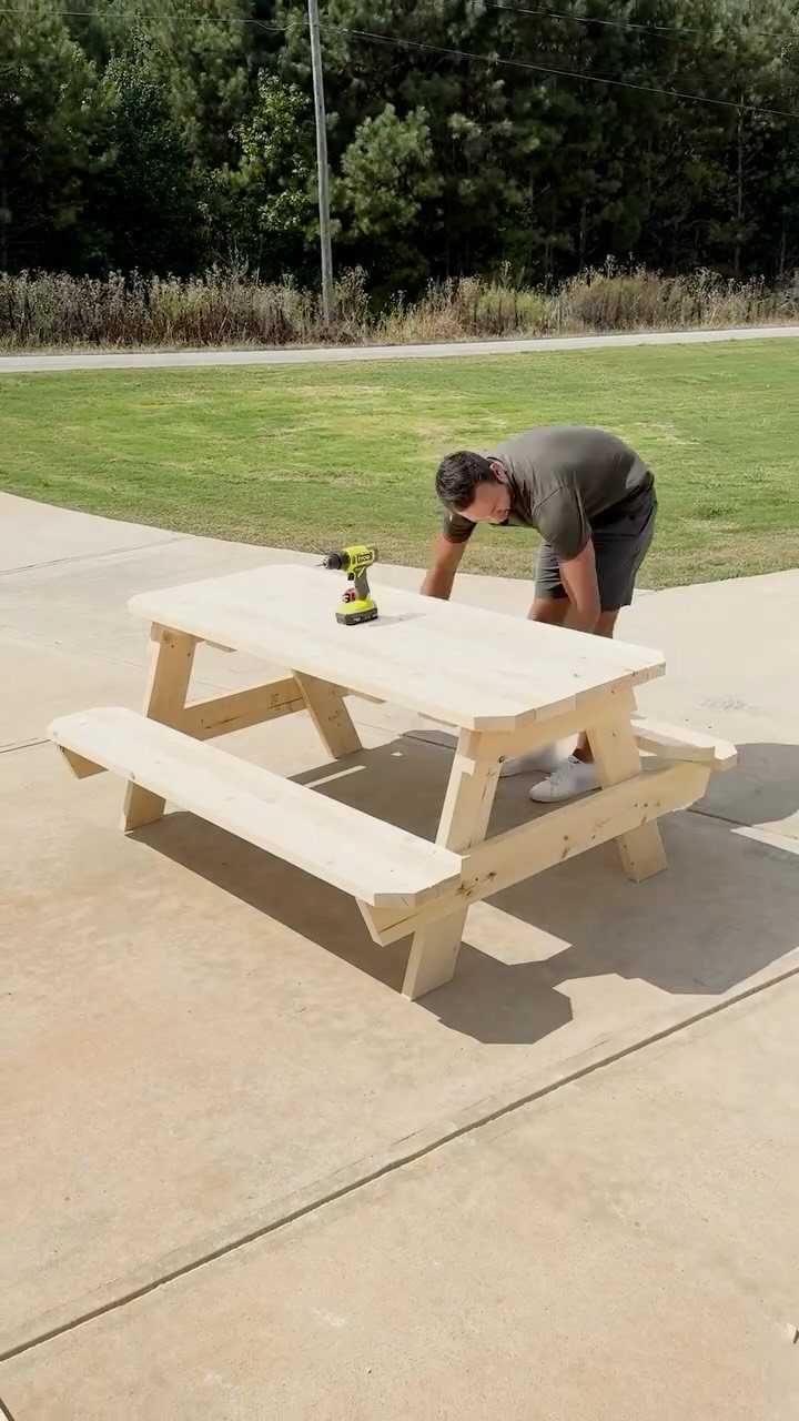 A man using a power drill to assemble a light wood picnic table outdoors.
