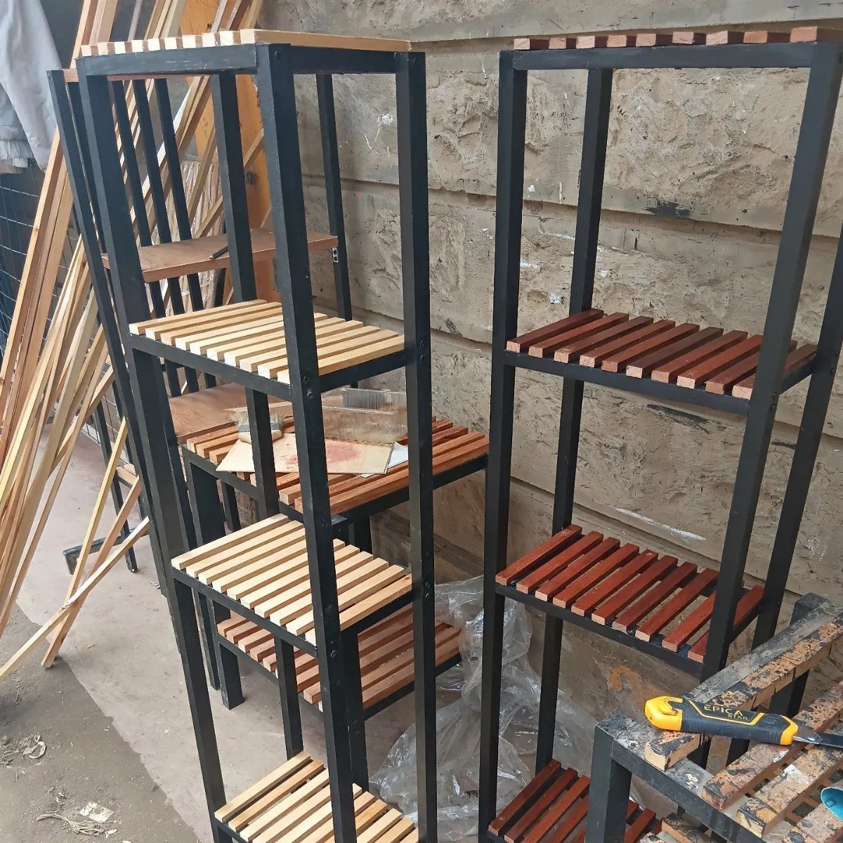 Two tall black metal shelving units with wooden slats standing against a stone wall.