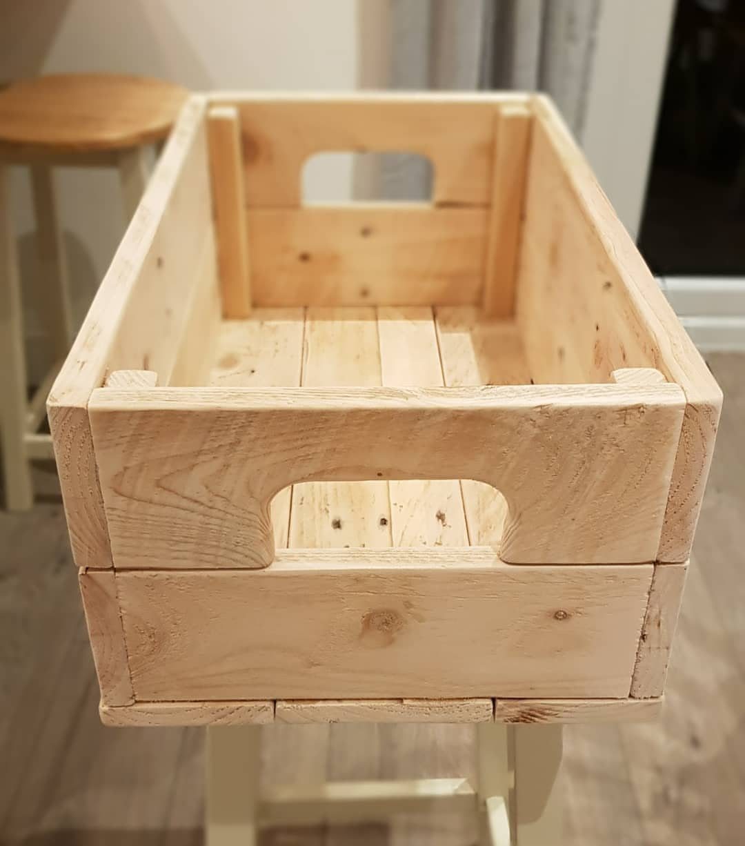 A light-colored wooden crate with cutout handles sitting on a white stool.